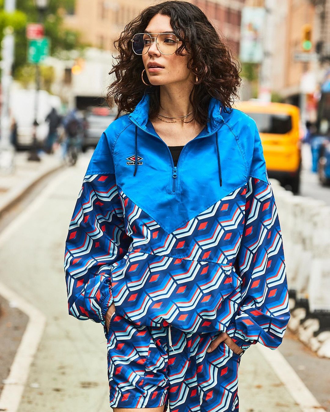Woman in patterned blue Umbro tracksuit poses on city street with taxi. Fashion shot with urban background and natural lighting.