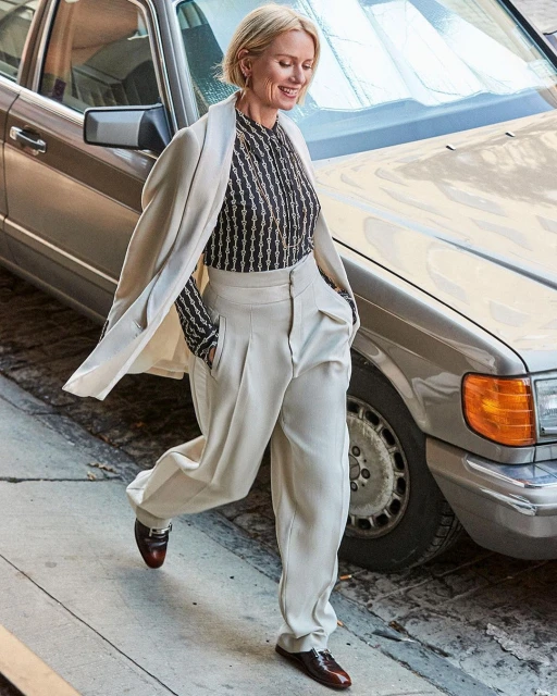 Naomi Watts in a patterned blouse and beige wide-leg trousers, walking on a sidewalk next to a silver vintage Mercedes-Benz.