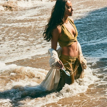 Woman in an olive green wrap top and trousers standing waist-deep in ocean waves, holding sunglasses