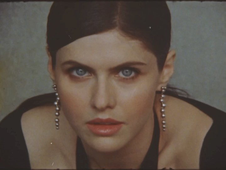 Close-up portrait of a Caucasian woman with dark hair and light eyes, wearing dangling earrings, against a muted background.