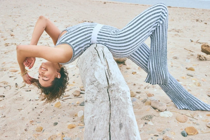 A woman in a jumpsuit balances on a driftwood log on a sandy beach, smiling with her head upside down in a playful pose.