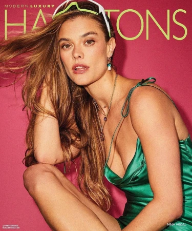 Close-up portrait of a woman with long brown hair wearing a green silk dress and white sunglasses on her head against a pink backdrop