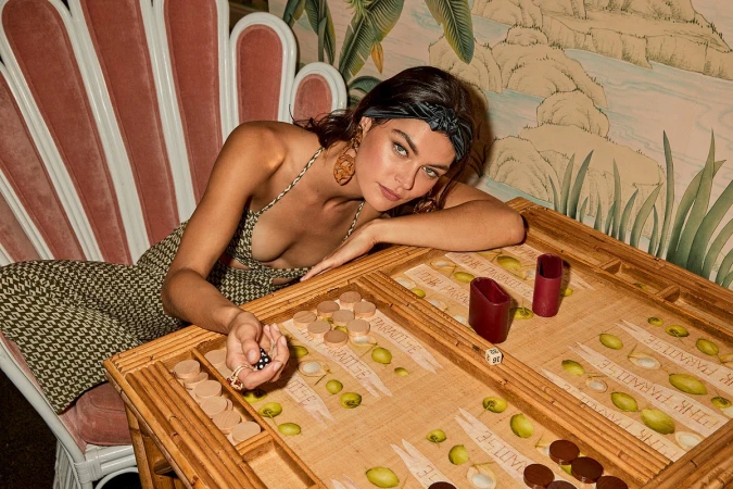 Woman playing backgammon at a table in an indoor setting with a pink shell chair and tropical wallpaper. The focus is on her hand with dice.