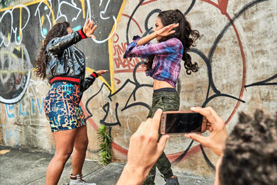 Two young women pose for a mobile phone photo in front of urban graffiti art with concrete; hands held up and casual trendy clothing.