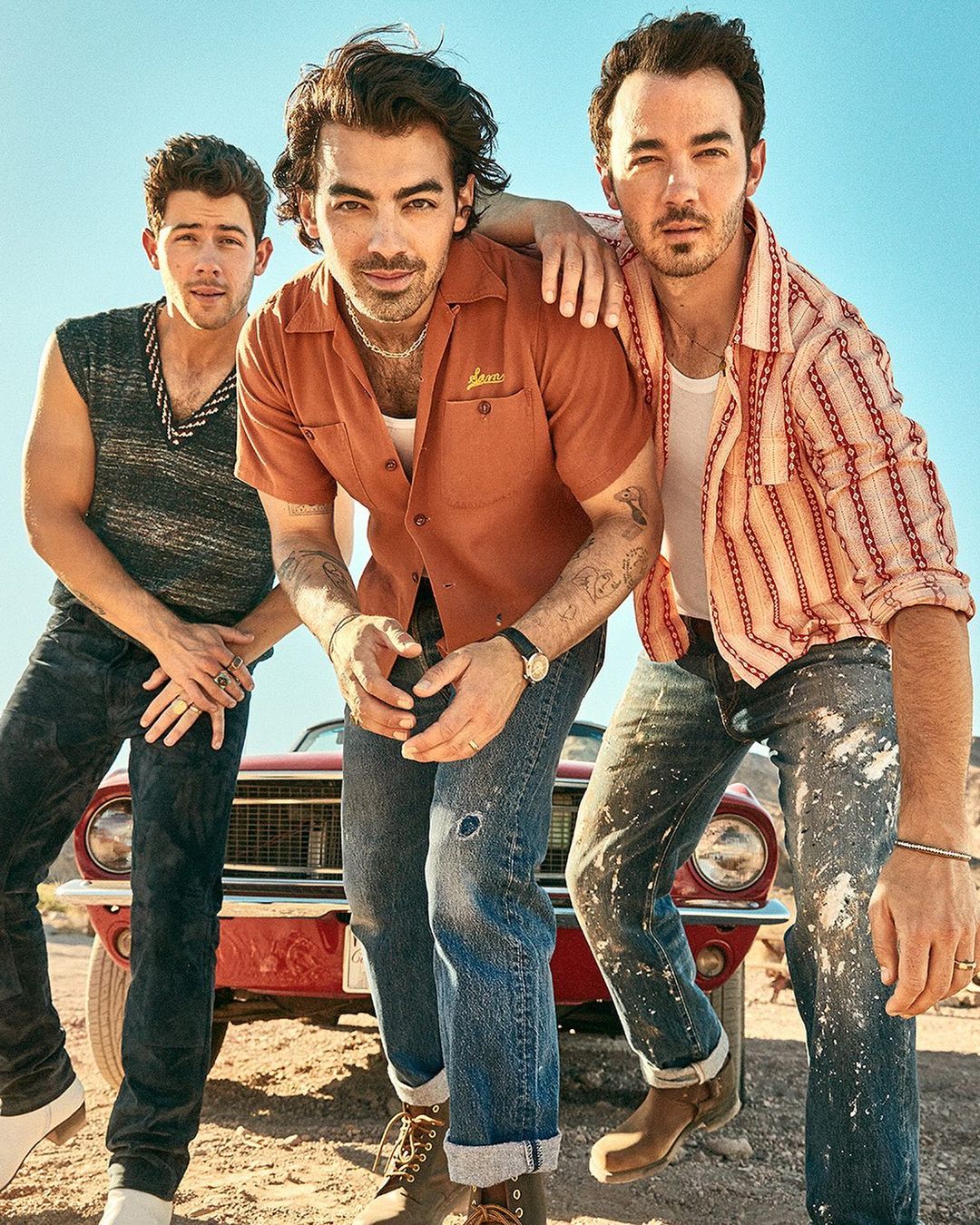 Three adult men pose outdoors with a red vintage car; the men wear casual clothing and the sky is light blue.