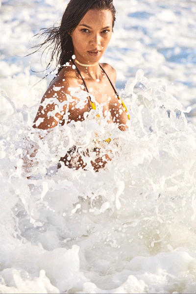 Adult female in a yellow bikini stands in ocean waves, looking toward the viewer on a sunny day at the beach.