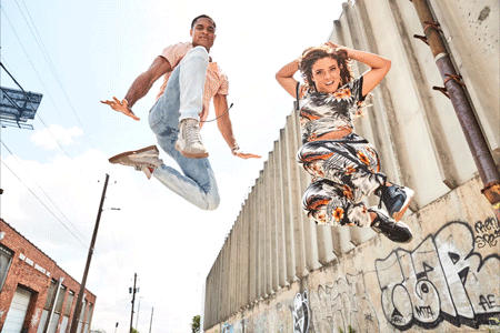 Two people jump in the air against an urban wall. The male wears jeans and a peach shirt while the female has patterned pants and top.