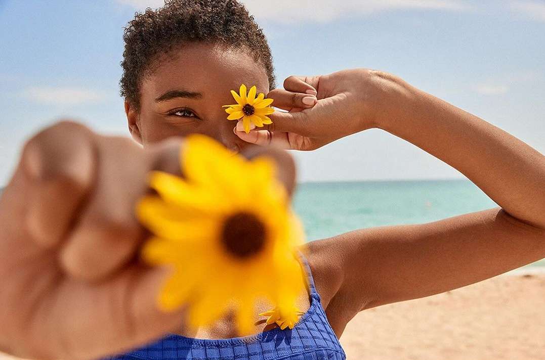 Close-up of a smiling, young, adult woman holding yellow sunflowers with a beach and ocean backdrop, creating a serene summer vibe.