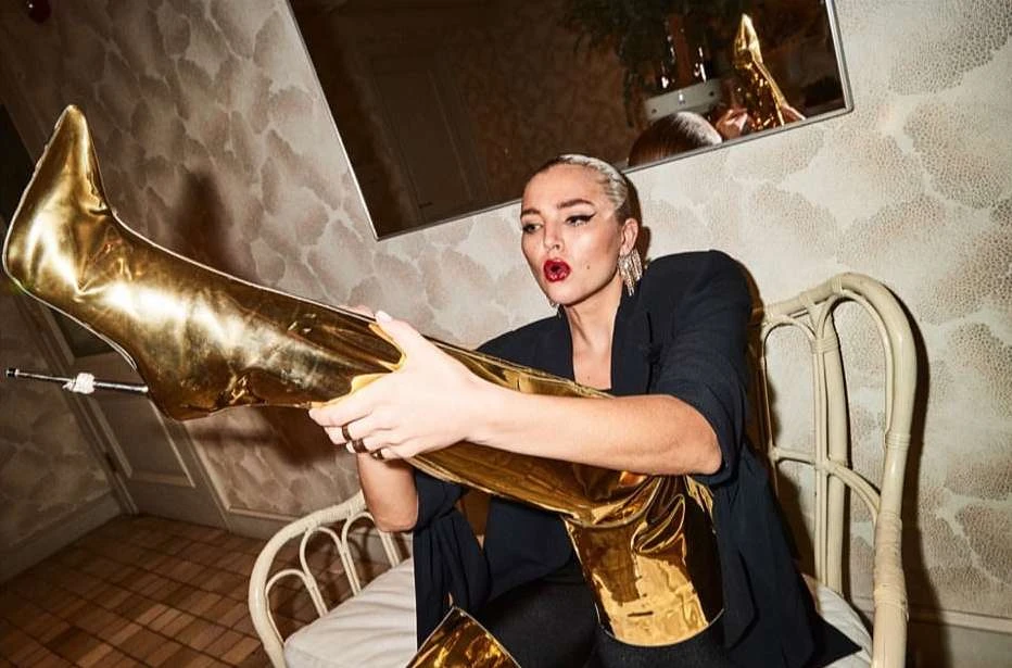 Blonde woman in a black jacket sitting on a white chair holding up a gold thigh high heel boot in front of a patterned wall.