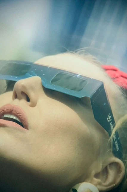 Close-up of a blond woman wearing eclipse glasses looking up at the sky. Her face is illuminated by bright, diffused sunlight.