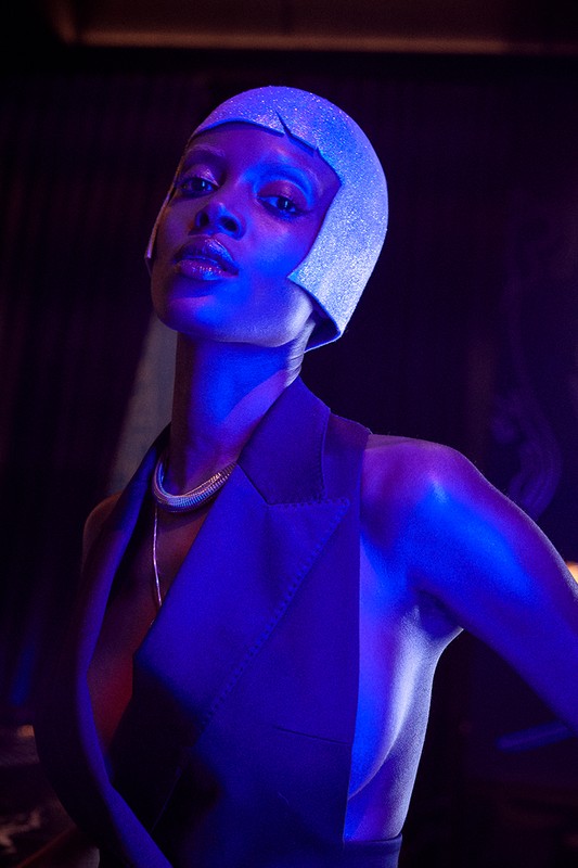 Close-up portrait of a stylish African American woman wearing a glittery cloche hat and sleeveless black jacket in blue lighting.