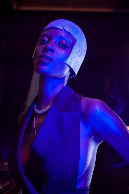 Close-up portrait of a stylish African American woman wearing a glittery cloche hat and sleeveless black jacket in blue lighting.