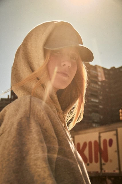 Young woman with auburn hair wearing a hoodie and baseball cap, poses in an urban environment with city buildings visible in background.