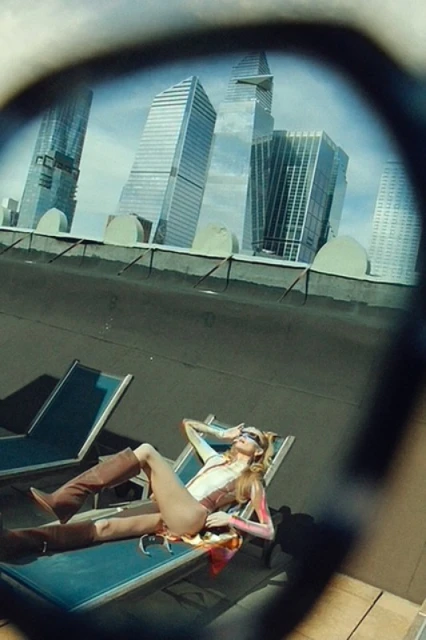 Woman in bodysuit and boots lounges on a deck chair on a rooftop, with skyscrapers visible in the background. Sunglasses reflection.