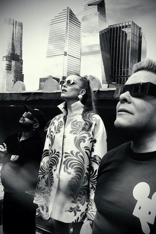 Three people in sunglasses gaze up at skyscrapers; a black and white photo captures a group against a modern urban backdrop.