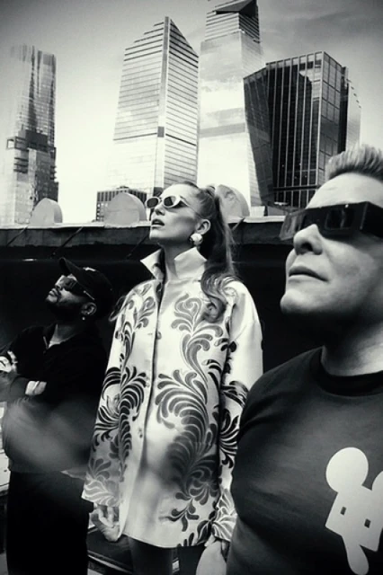 Three people in sunglasses gaze up at skyscrapers; a black and white photo captures a group against a modern urban backdrop.