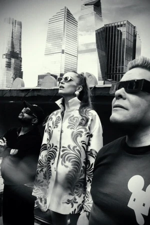 Three people in sunglasses gaze up at skyscrapers; a black and white photo captures a group against a modern urban backdrop.