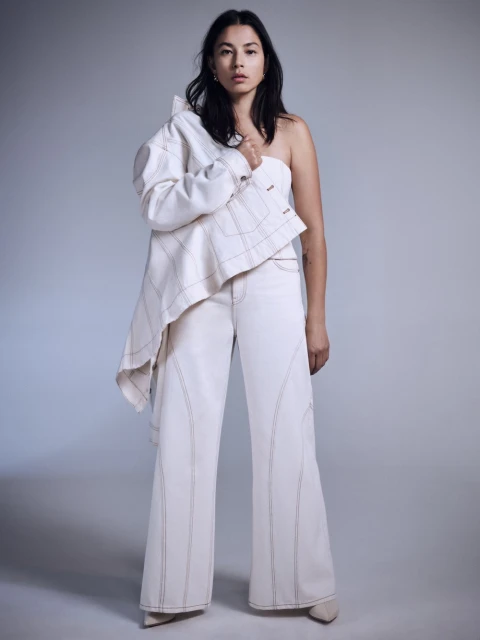 A woman with dark hair poses in a studio wearing white wide-leg jeans and a white top, with a jacket over her shoulder.