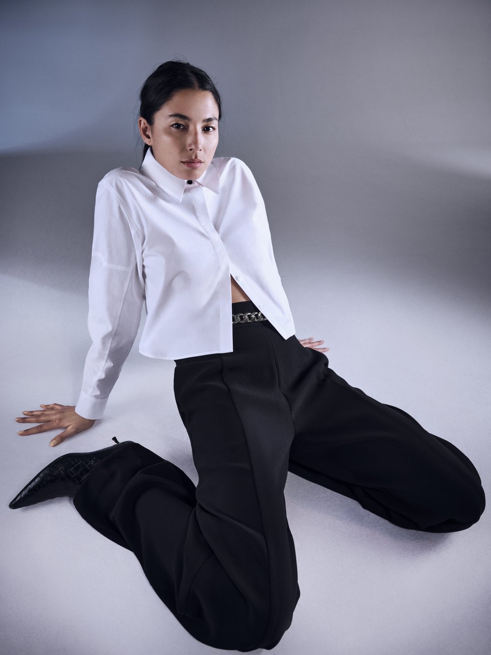 Elegant woman with dark hair poses in a white button-down shirt, black pants, and pointed boots against a neutral studio backdrop.