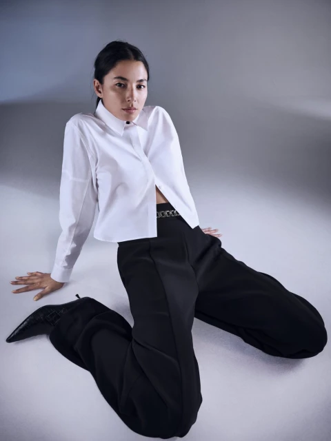 Elegant woman with dark hair poses in a white button-down shirt, black pants, and pointed boots against a neutral studio backdrop.