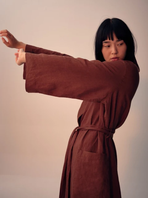 Young woman in a brown robe-style dress poses with outstretched arms against a neutral background in a studio setting.