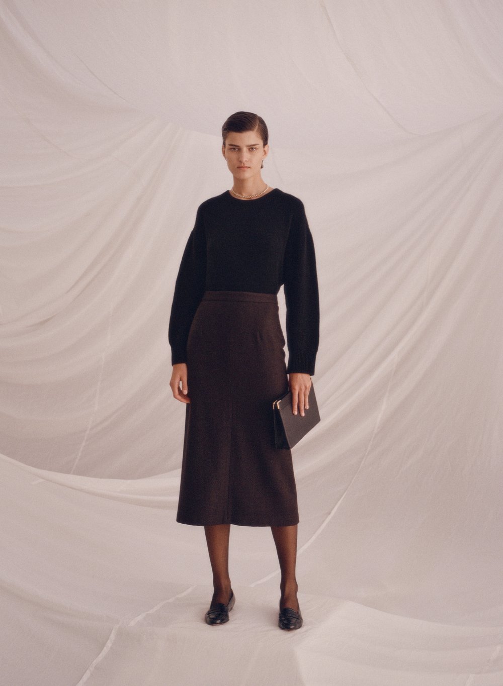 Full-shot of a woman in a black sweater and brown skirt holding a clutch, standing against a white draped background in a fashion pose.