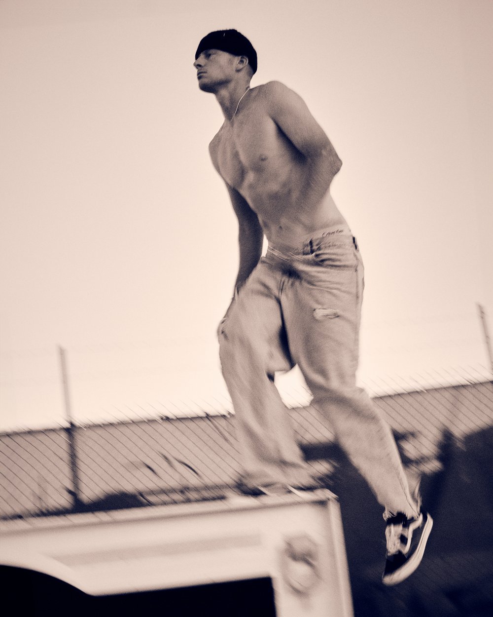 Athletic man in motion captured mid-jump in monochrome, wearing casual pants and sneakers against a bright sky and urban backdrop.