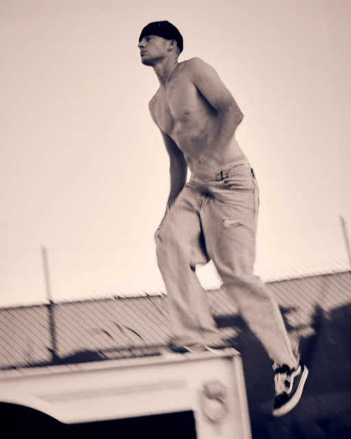 Athletic man in motion captured mid-jump in monochrome, wearing casual pants and sneakers against a bright sky and urban backdrop.
