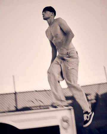 Athletic man in motion captured mid-jump in monochrome, wearing casual pants and sneakers against a bright sky and urban backdrop.