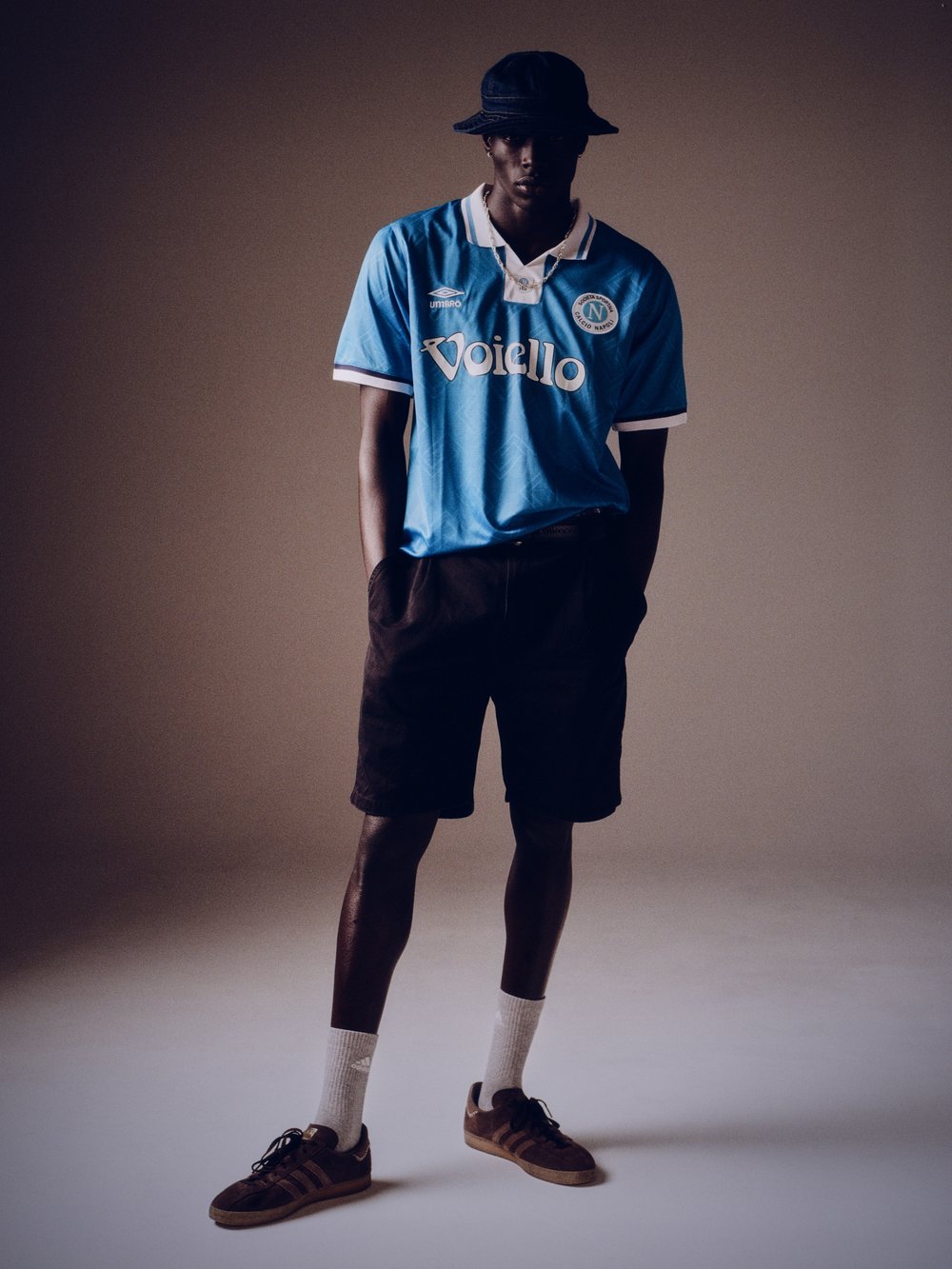 A dark-skinned male model poses indoors wearing a vintage soccer jersey, dark shorts, socks, Adidas Sambas, and a matching bucket hat.