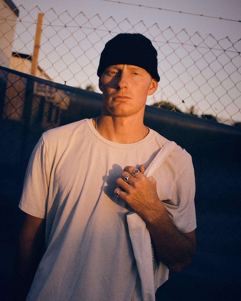 Portrait of a man wearing a black beanie and white t-shirt standing outdoors against a chain-link fence during golden hour sunset.