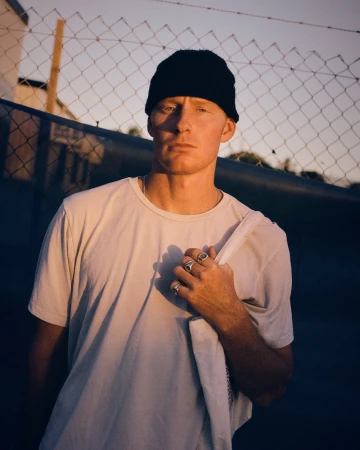 Portrait of a man wearing a black beanie and white t-shirt standing outdoors against a chain-link fence during golden hour sunset.