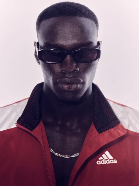 Athletic dark-skinned man in sunglasses and red Adidas track jacket poses against a white backdrop in a studio environment.