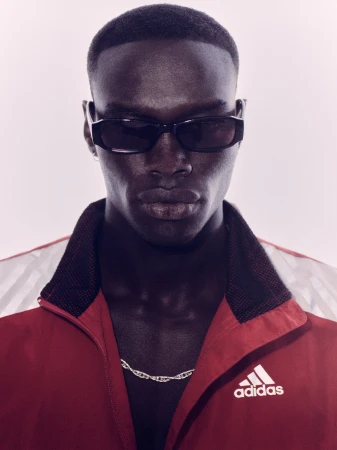 Close-up portrait of a Black man wearing dark sunglasses and a red zip jacket with a silver chain necklace against a light background.