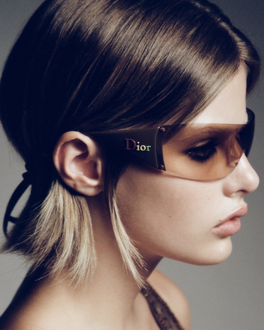 A close-up studio shot captures a model with short brown hair and tinted sunglasses, emphasizing fashion and beauty in a neutral setting.