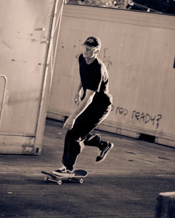 Skater in dark clothes and cap riding a skateboard outdoors on concrete by Lewis Stevenson.