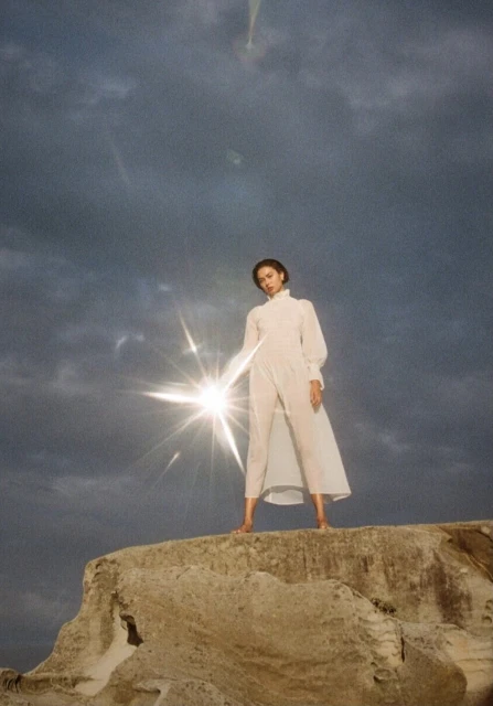 Woman in a white jumpsuit stands on a rocky surface with a cloudy sky, illuminated by a bright light shining towards the camera.