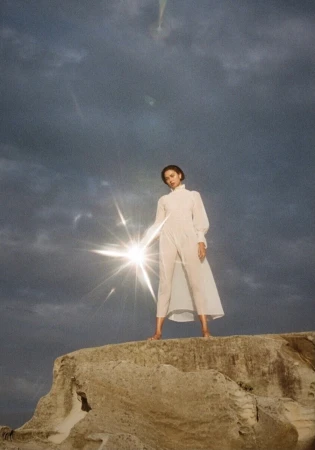 Woman in a white jumpsuit stands on a rocky surface with a cloudy sky, illuminated by a bright light shining towards the camera.