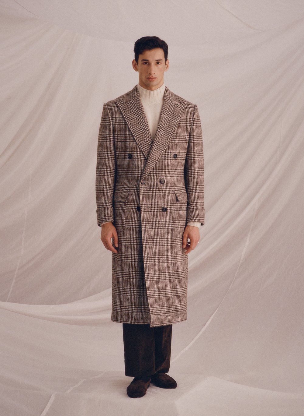 A stylish man stands in a full-length shot wearing a brown plaid overcoat, white turtleneck, and dark brown trousers against a draped background.