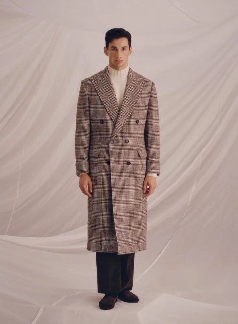 A stylish man stands in a full-length shot wearing a brown plaid overcoat, white turtleneck, and dark brown trousers against a draped background.