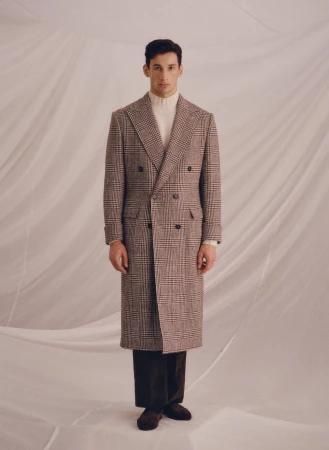 A stylish man stands in a full-length shot wearing a brown plaid overcoat, white turtleneck, and dark brown trousers against a draped background.
