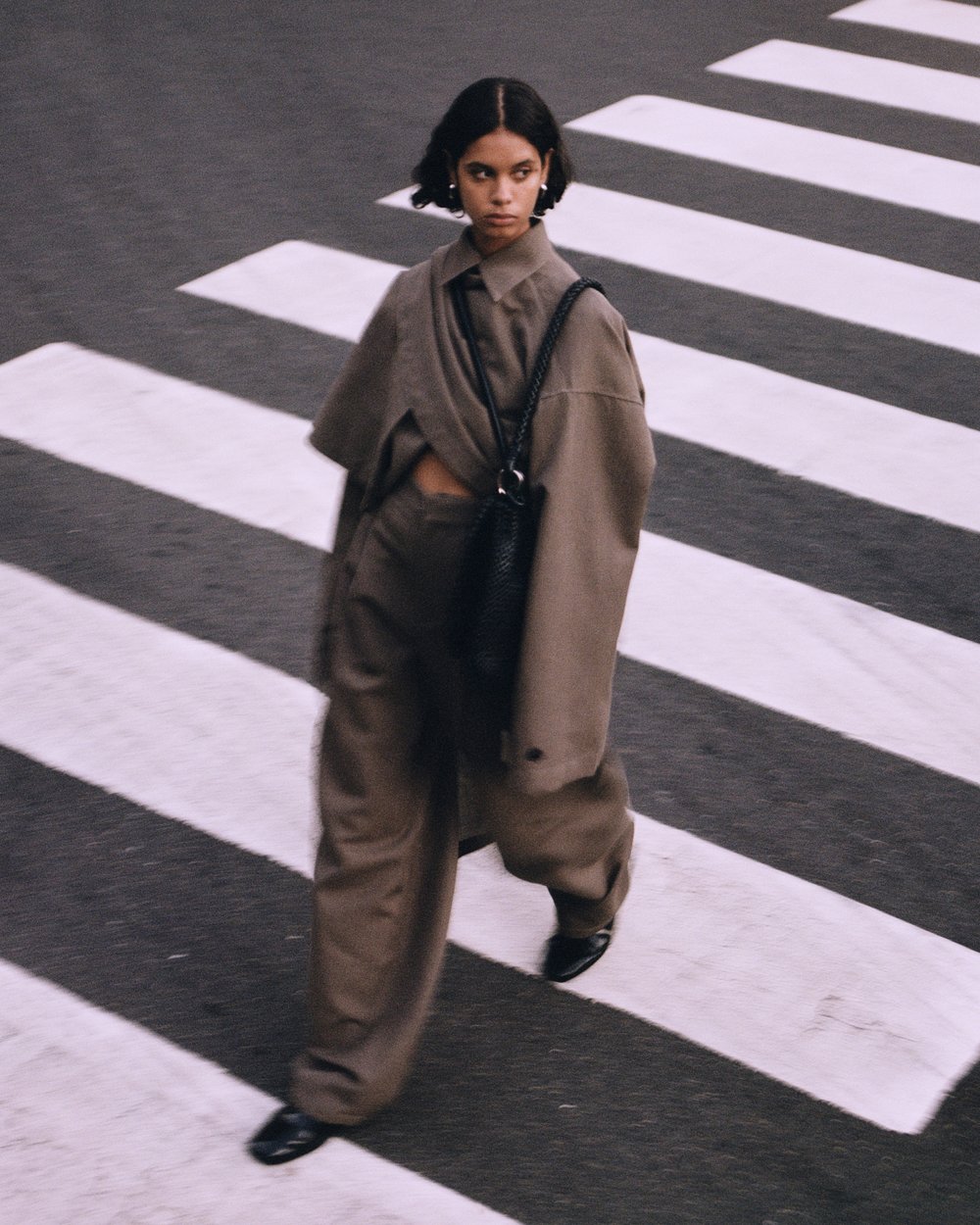 A woman in a monochrome taupe outfit walks across a pedestrian crosswalk, glancing to the side with a slight smile.
