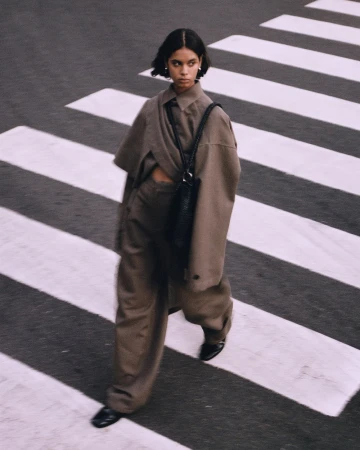 A woman in a monochrome taupe outfit walks across a pedestrian crosswalk, glancing to the side with a slight smile.