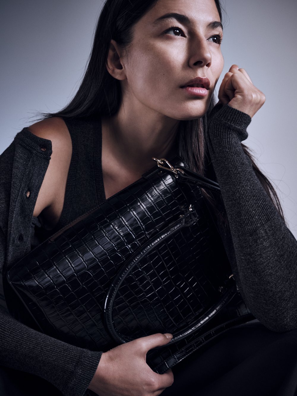 Portrait of an adult female holding a black, textured handbag with a grey cardigan and black top, set against a neutral background.