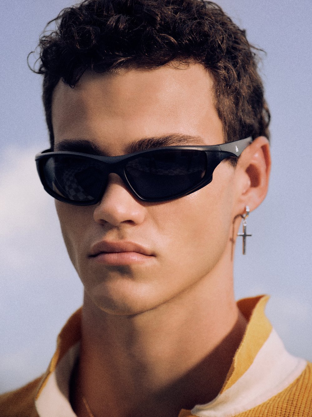 Close-up shot of a young adult male wearing sunglasses, a yellow polo shirt, and a silver cross earring against a blue sky backdrop.