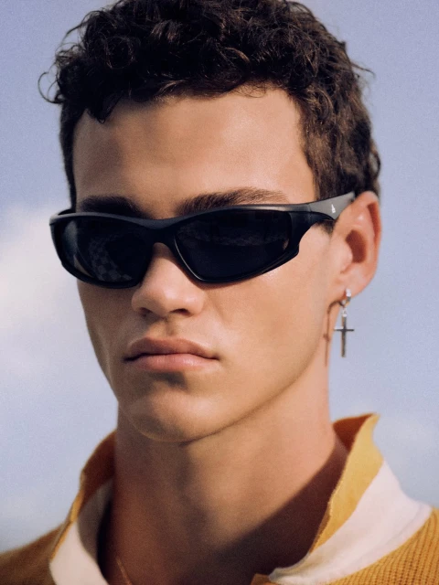 Close-up shot of a young adult male wearing sunglasses, a yellow polo shirt, and a silver cross earring against a blue sky backdrop.