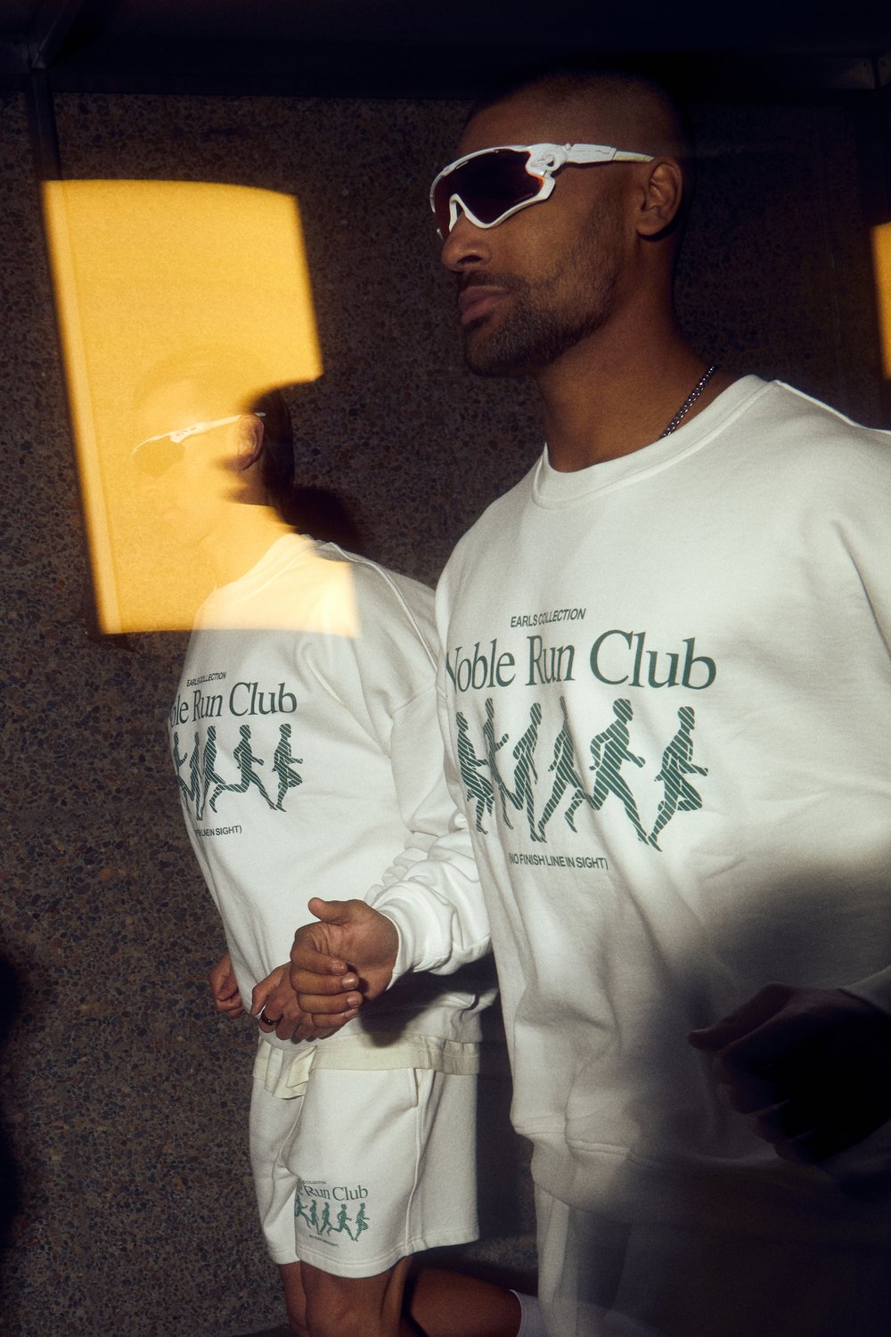 Two adult males pose indoors in off-white athletic apparel with green graphics and sunglasses. Selective focus and bright lighting.