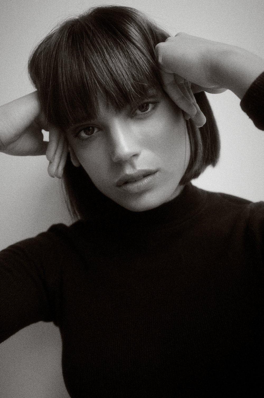 Monochrome portrait of a young adult woman with hands near her head, wearing a dark turtleneck, looking directly at the camera.