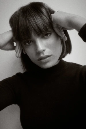 Monochrome portrait of a woman with a blunt bob haircut and bangs, wearing a dark turtleneck, posing with hands near her temples.