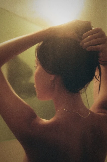 Adult female in warm-toned indoor setting, touches her hair. Gold necklace and hoop earrings are visible in a candid shot.